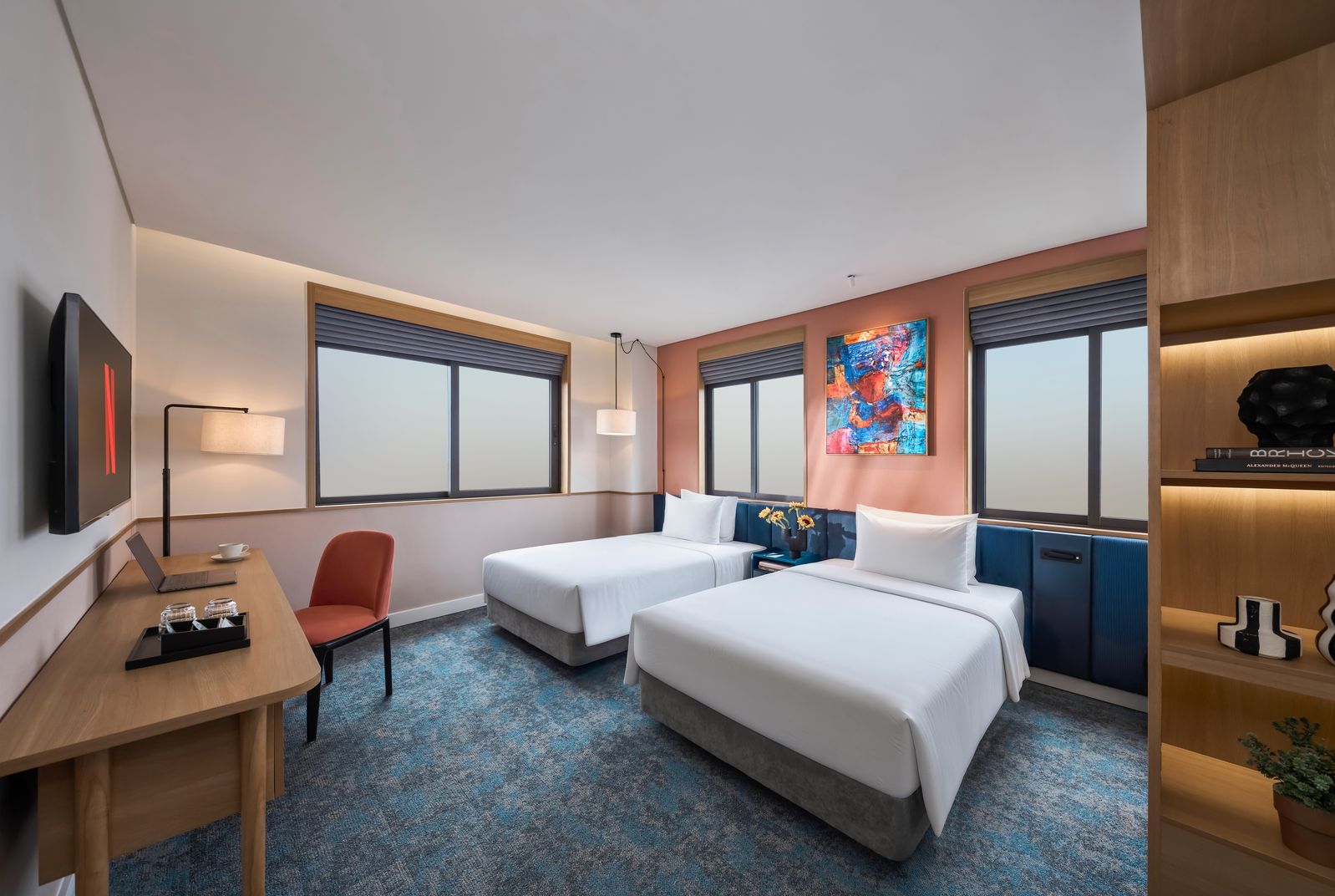 A hotel room with two beds, a desk with a laptop, a chair, a TV, and a large window.