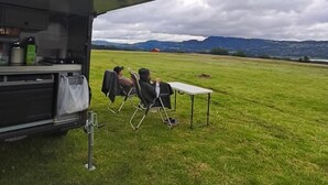Montaje para acampar con una mesa y sillas en un campo cubierto de hierba, un vehículo con compartimentos de almacenamiento y montañas al fondo.