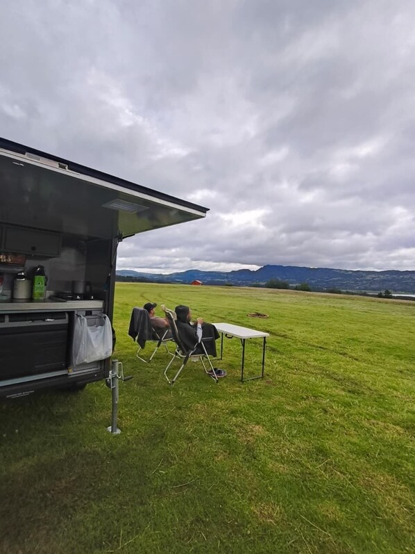 A camping setup with a table and chairs in a grassy field, a vehicle with storage compartments, and a mountainous backdrop. - Camping San Francisco - Cabaña, Teepee & Zona de Camping (Guatavita)