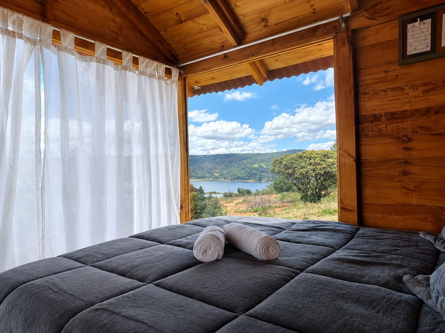 Un letto con una coperta grigia e asciugamani arrotolati, un soffitto in legno e una finestra vista lago e colline.