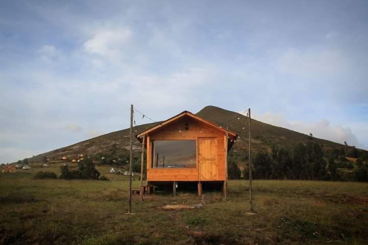 Una cabaña de madera con un ventanal grande, ubicada en un campo con una colina al fondo.