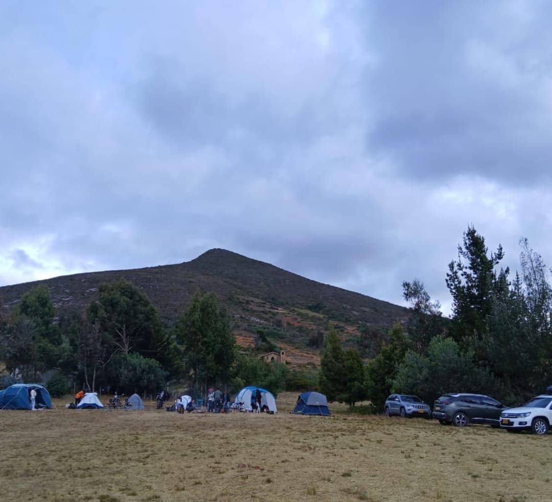 Un sitio de camping con tiendas de campaña y coches, una colina al fondo y el texto "Guatavita, Cund".