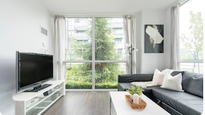 Un salon moderne doté d’un téléviseur à écran plat, d’un canapé foncé, d’une table basse blanche et d’une grande fenêtre offrant une vue sur la verdure et les bâtiments.