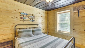 Camera da letto in una baita di legno con un letto, cuscini, una testiera in metallo, una finestra con persiane e una parete in legno con una freccia decorativa.