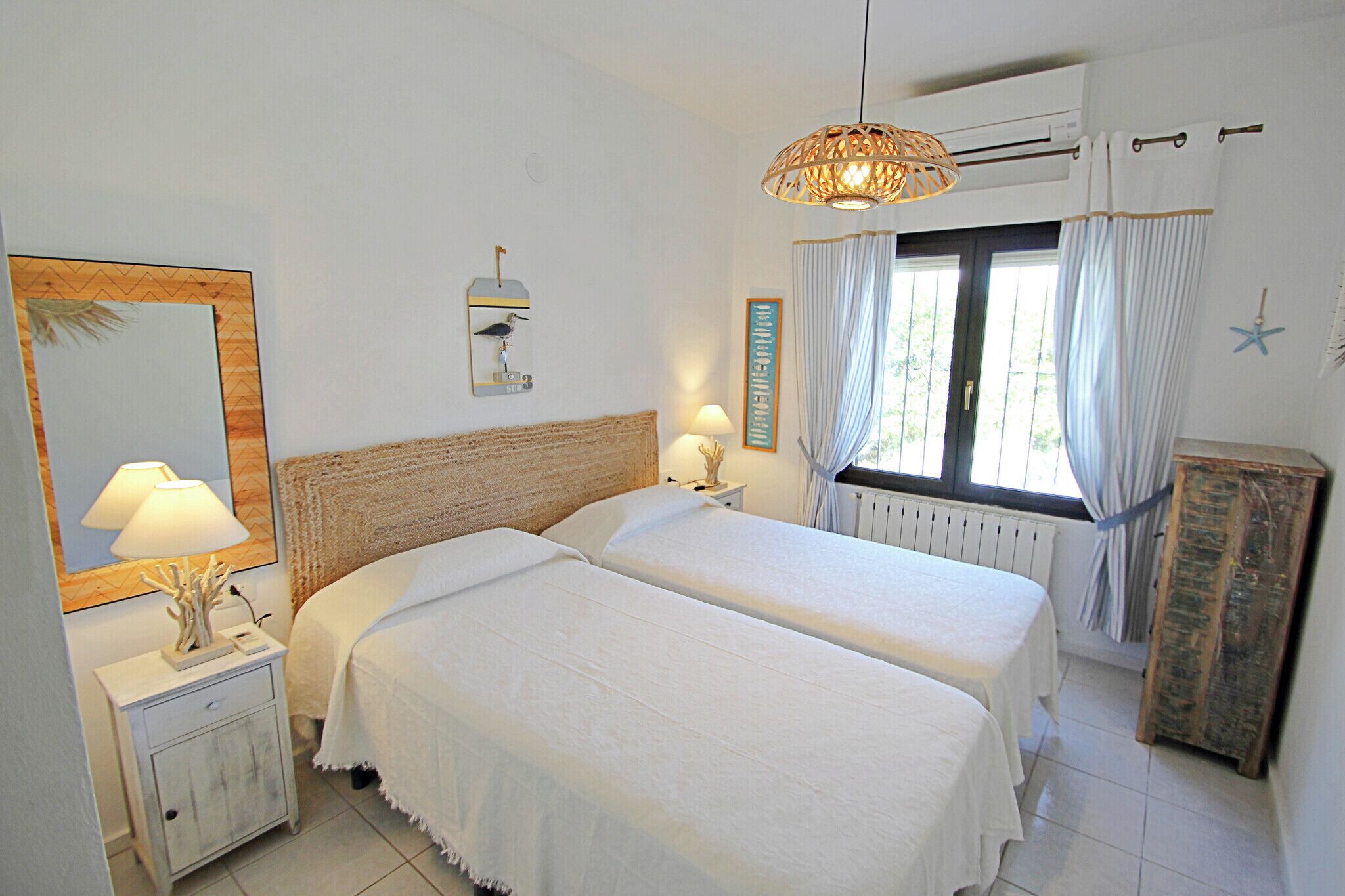 A bedroom with two beds, a nightstand with a lamp, a wall-mounted mirror, and a window with curtains.