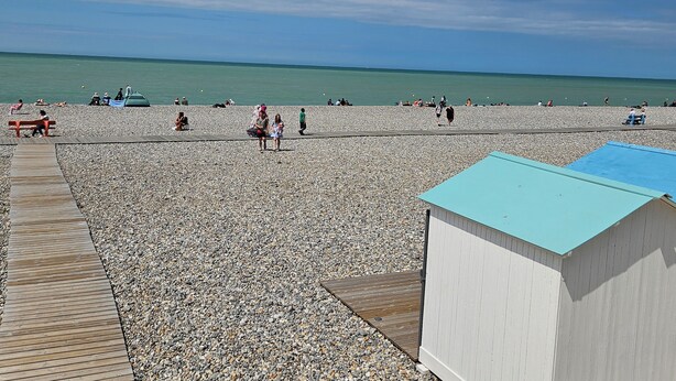Une plage de galets avec une promenade en bois et un petit bâtiment doté d’un toit bleu.