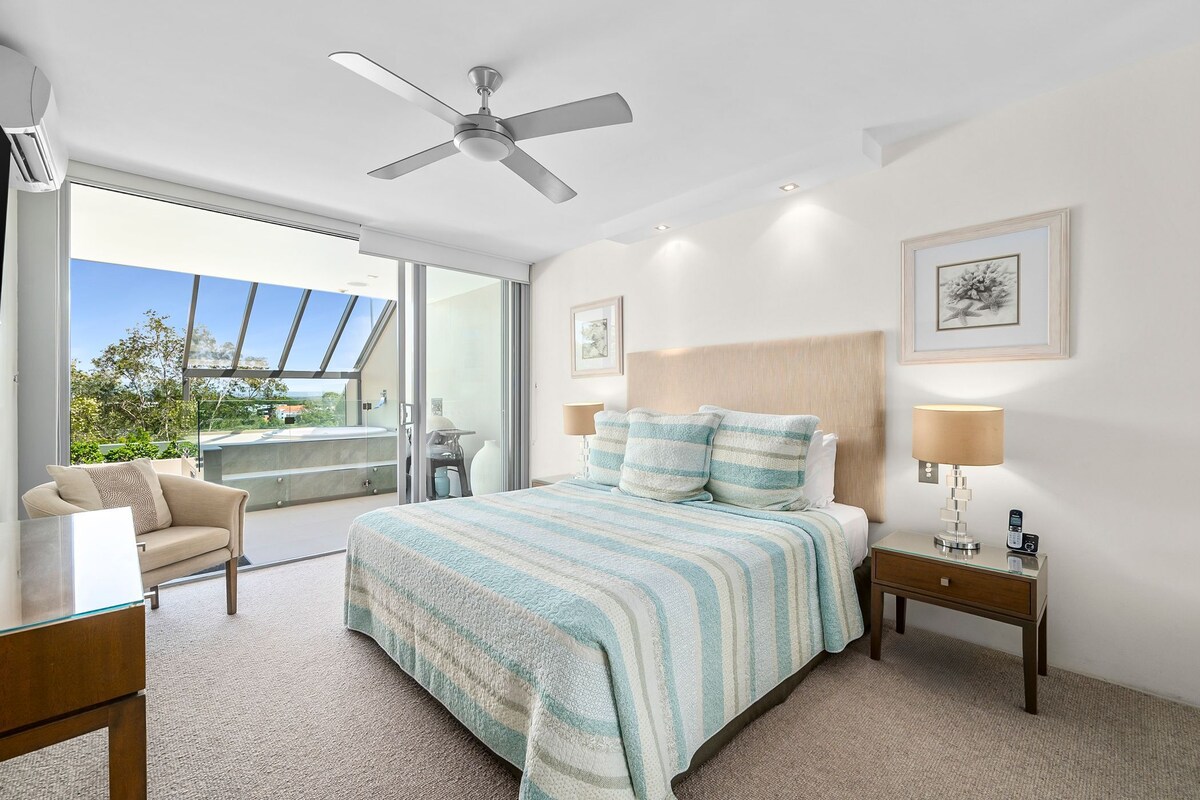 A bedroom with a bed, a nightstand, a chair, a ceiling fan, and a large window.