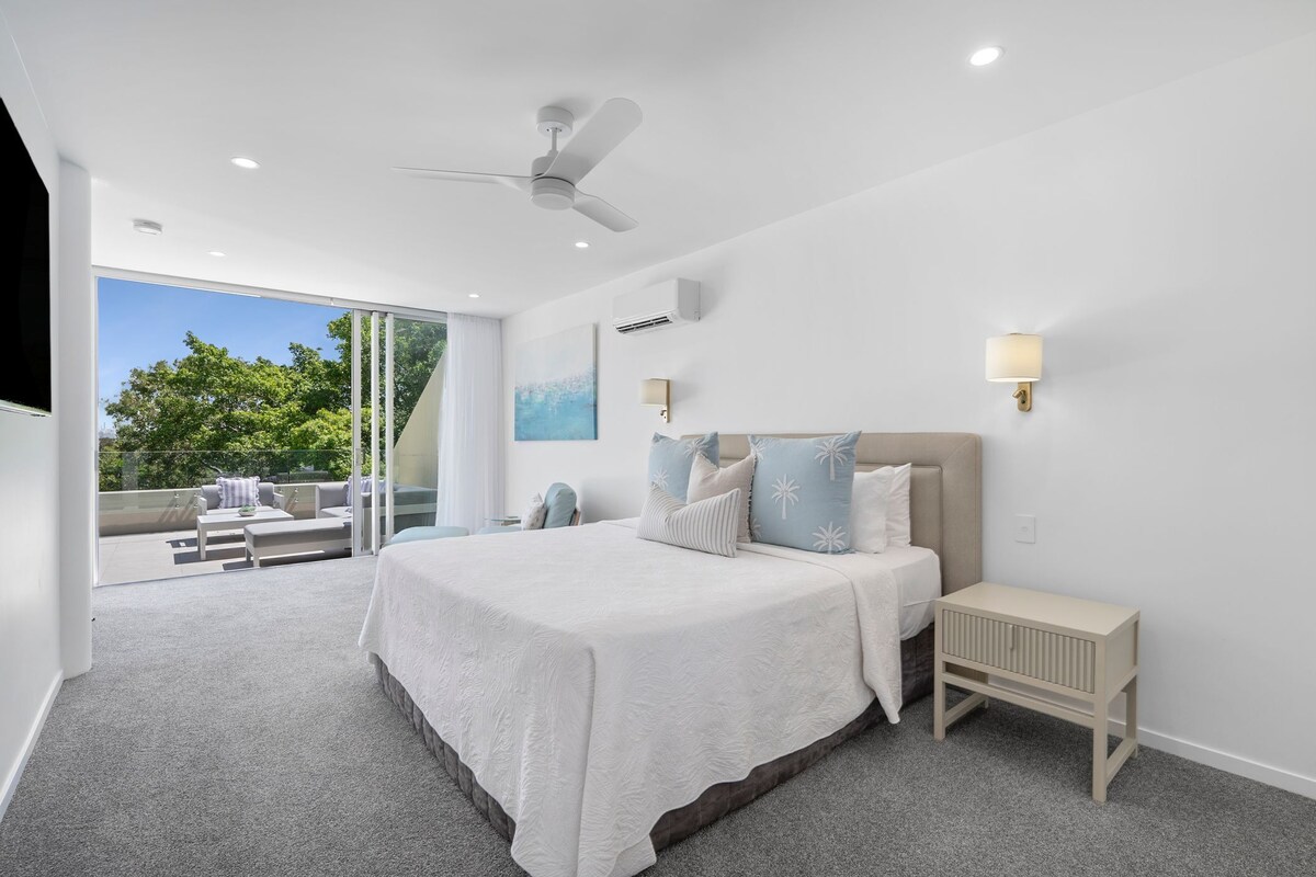 A modern bedroom with a large bed, a ceiling fan, a nightstand, and a view of the outdoors.