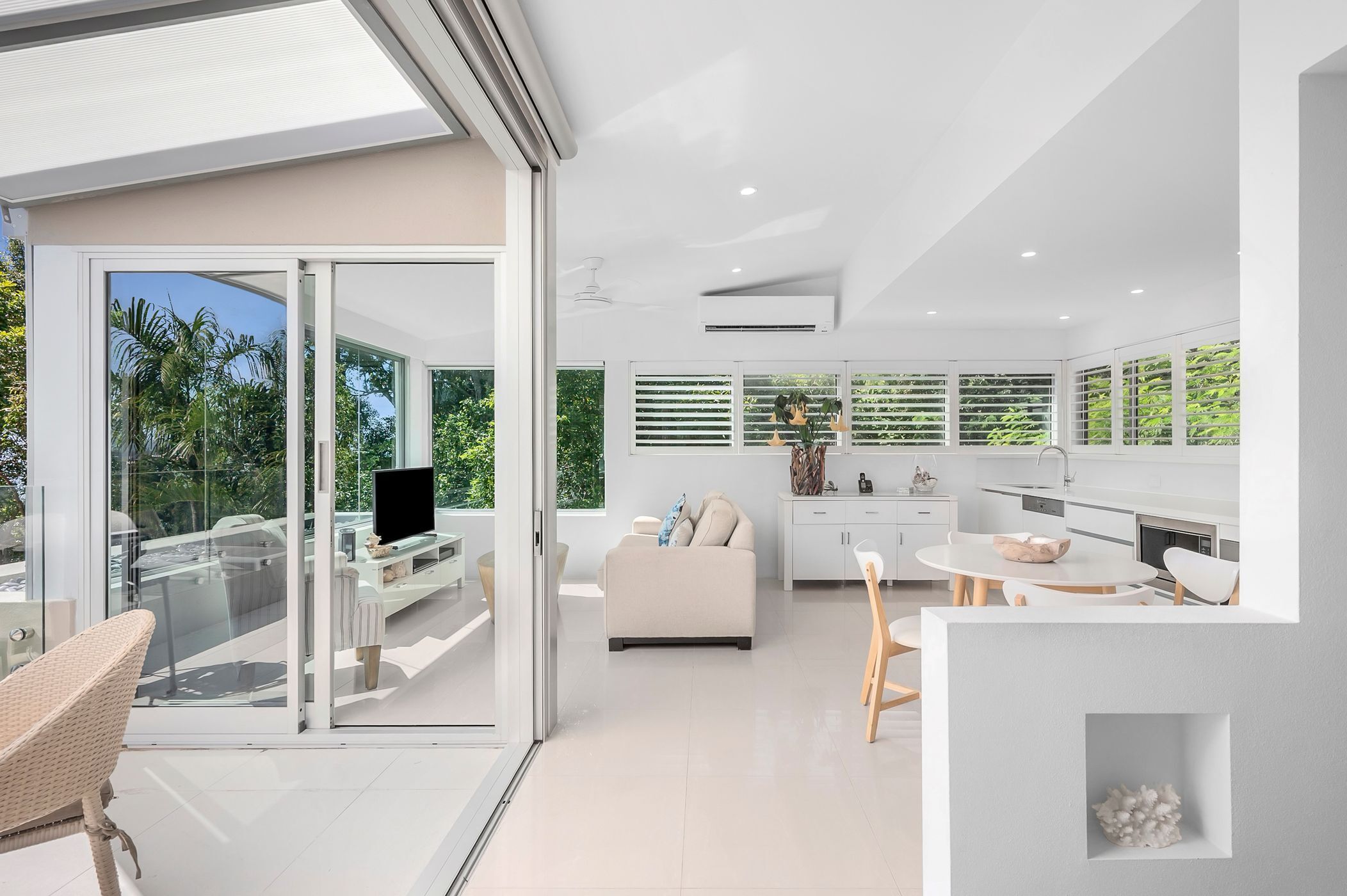 A modern interior with a glass sliding door leading to a balcony, a white sofa, a dining table with chairs, and a kitchen area with white cabinets and a built-in oven.