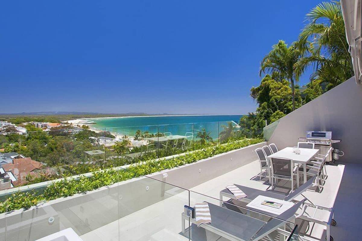 Une terrasse sur le toit dotée d’une balustrade en verre, d’un espace salon extérieur et offrant une vue sur l’océan et le paysage environnant.