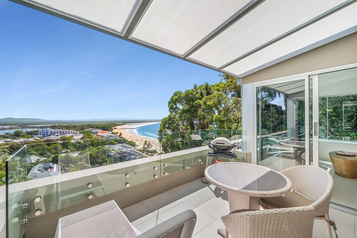 Un balcon doté d’une balustrade en verre, d’une table et de chaises, offrant une vue sur une plage et l’océan.