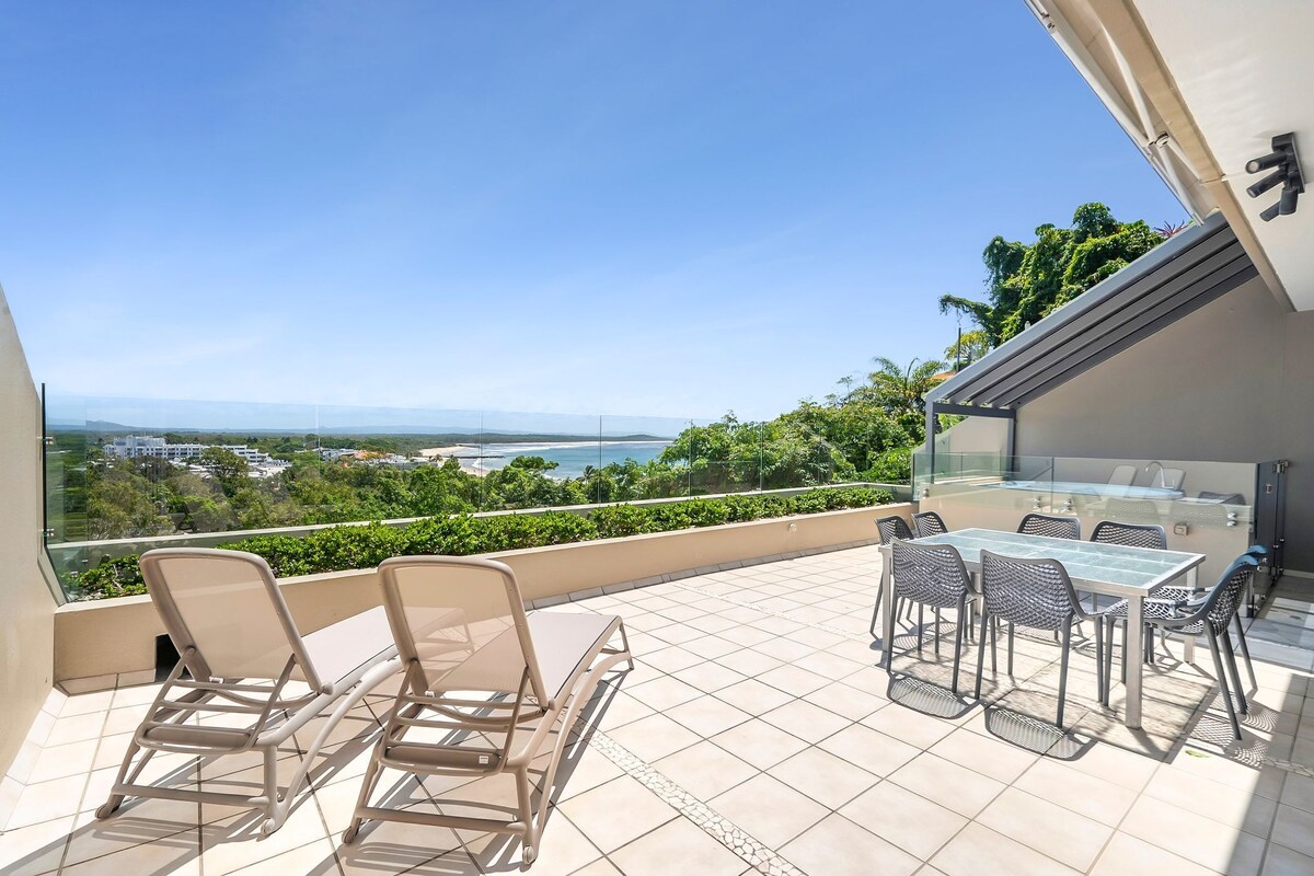 A terrace with a glass balustrade, outdoor seating, and a view of the sea and landscape.