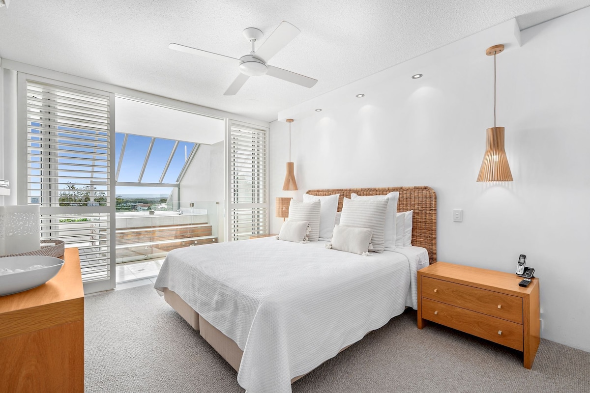 A modern bedroom with a large bed, a wooden nightstand, a ceiling fan, and a view through the glass door.
