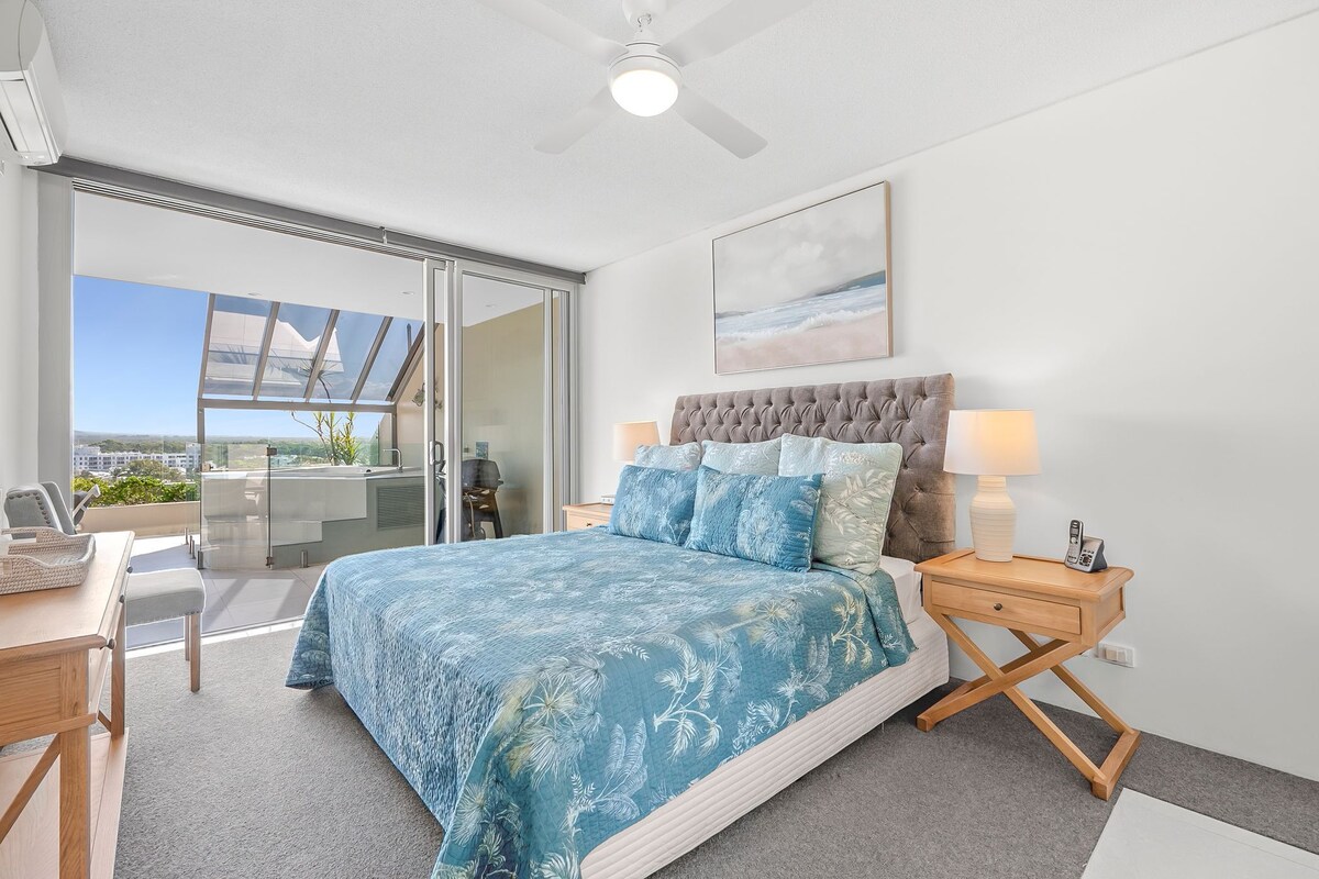 A bedroom with a bed, bedside lamps, a ceiling fan, a window, and a view of the outdoors.