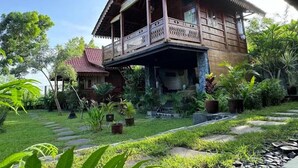 A traditional wooden house with a balcony, surrounded by lush greenery and stone pathways. - JavaLO Lite (Pujut)