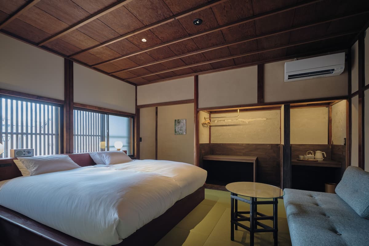A hotel room with a bed, a small table, a sofa, a desk, and an air conditioning unit.