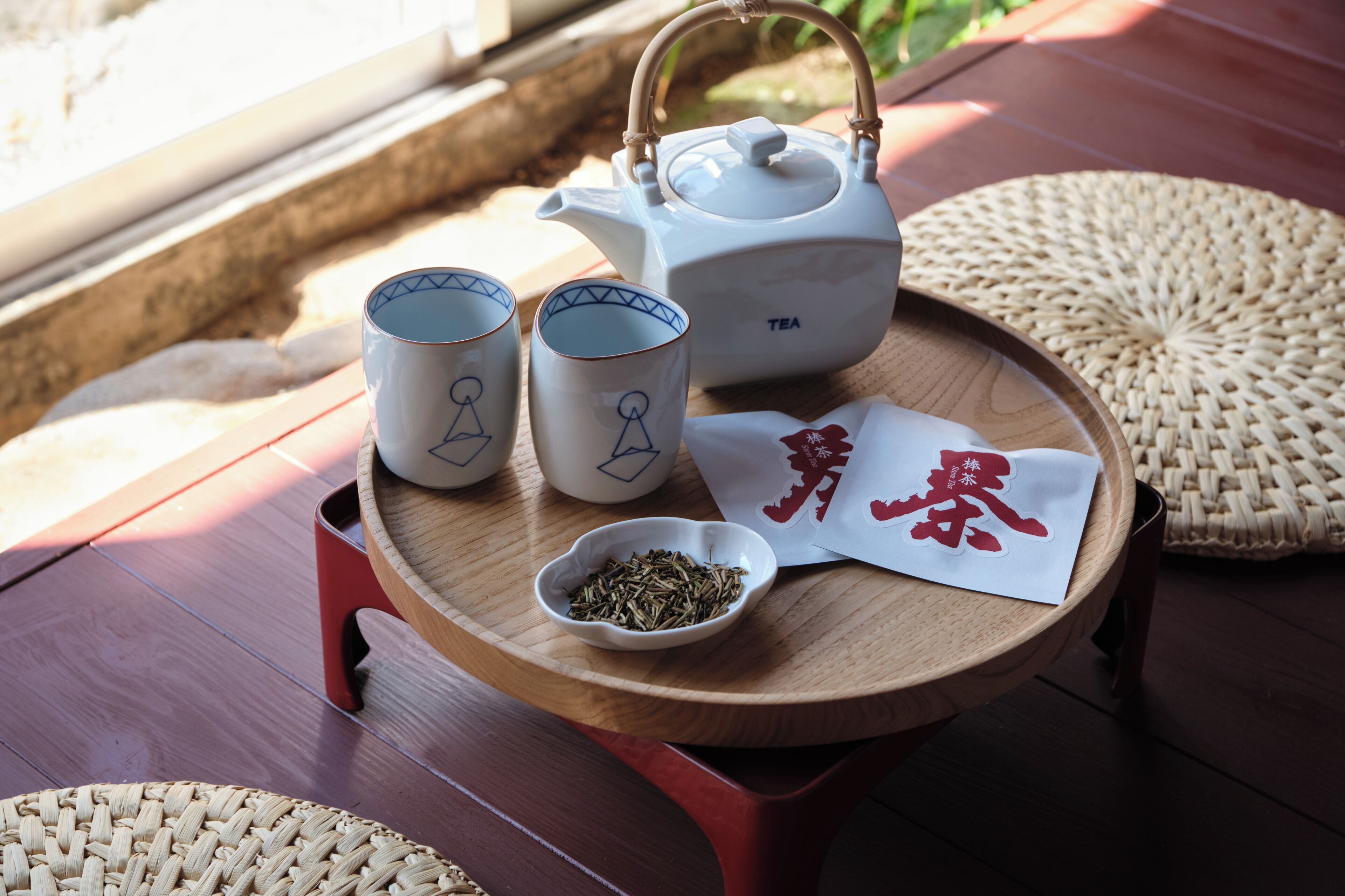 A wooden tray with a white teapot, two cups, a bowl of tea leaves, and a napkin with red text.