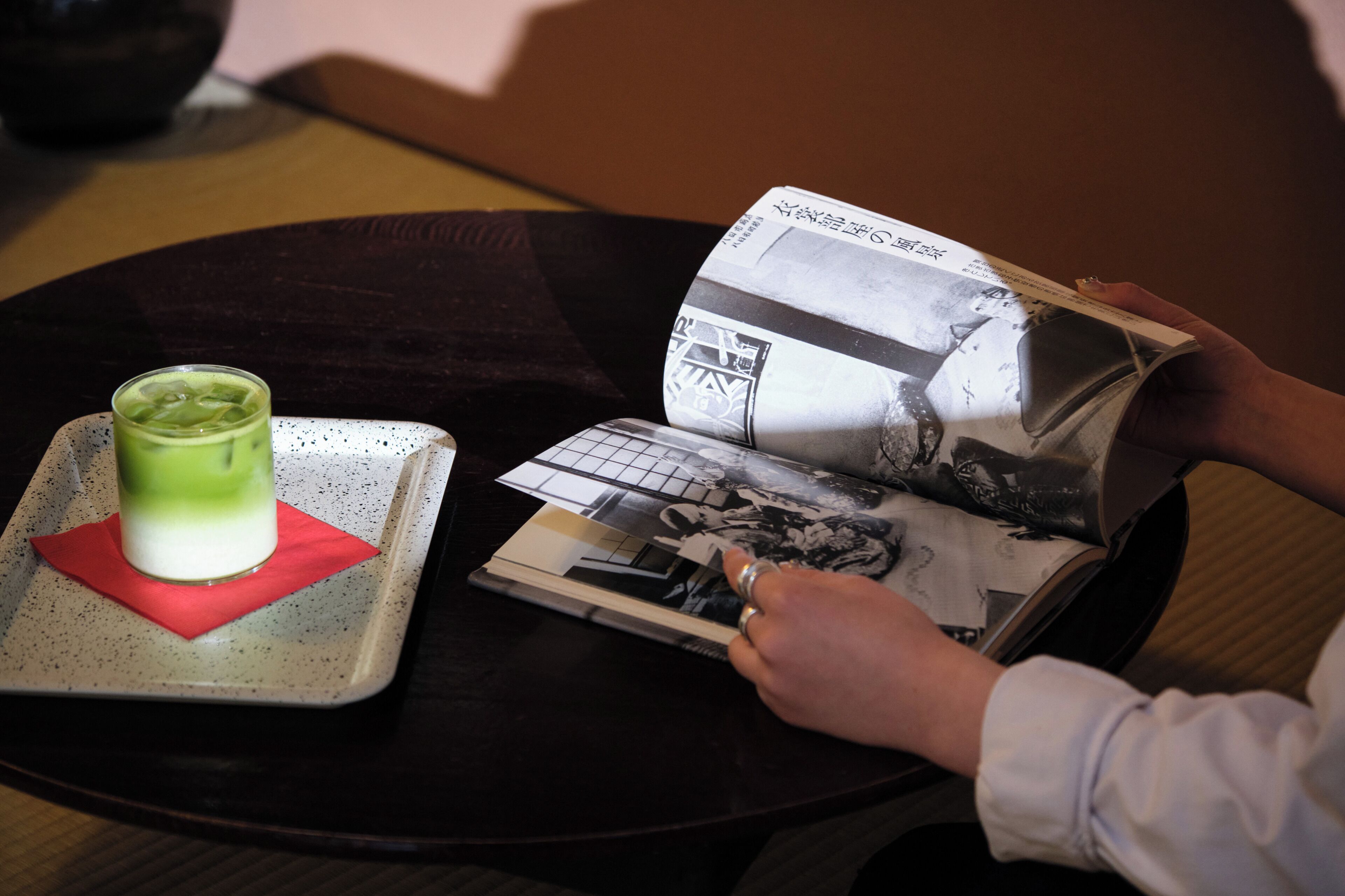 A person reading a magazine on a round table with a glass of green beverage and a red napkin.