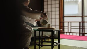 A person in traditional attire pouring tea into a cup on a small table in a room with tatami flooring and a sliding door. - Komado Bengara (Komatsu)