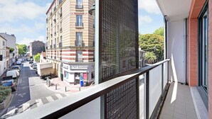 Blick von einem Balkon auf eine Straße mit parkenden Autos und Gebäuden, darunter ein mehrstöckiges Gebäude mit Balkonen und einer mit 'Parti Socialiste' beschrifteten Geschäftseröffnung.
