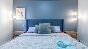 A neatly made bed with a geometric patterned bedspread, a blue headboard, and a framed picture on the wall.