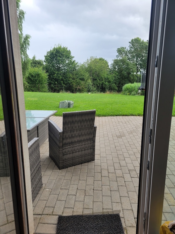 An outdoor patio with wicker furniture, a glass table, and a view of a green lawn and trees.