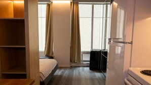 Een kleine kamer met een bed, een tafeltje, een raam met gordijnen, een radiator en een wit apparaat.