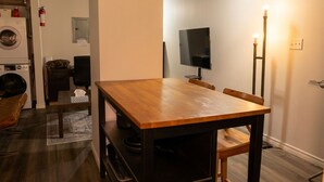 Een kamer met een houten eettafel, een flatscreen televisie, een staande vloerlamp en een wasmachine.
