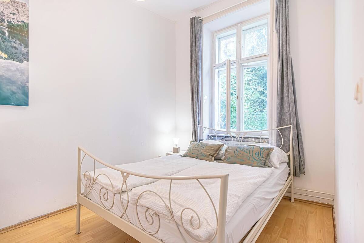 A bedroom with a metal bed frame, white bedding, a window with curtains, and a painting on the wall.