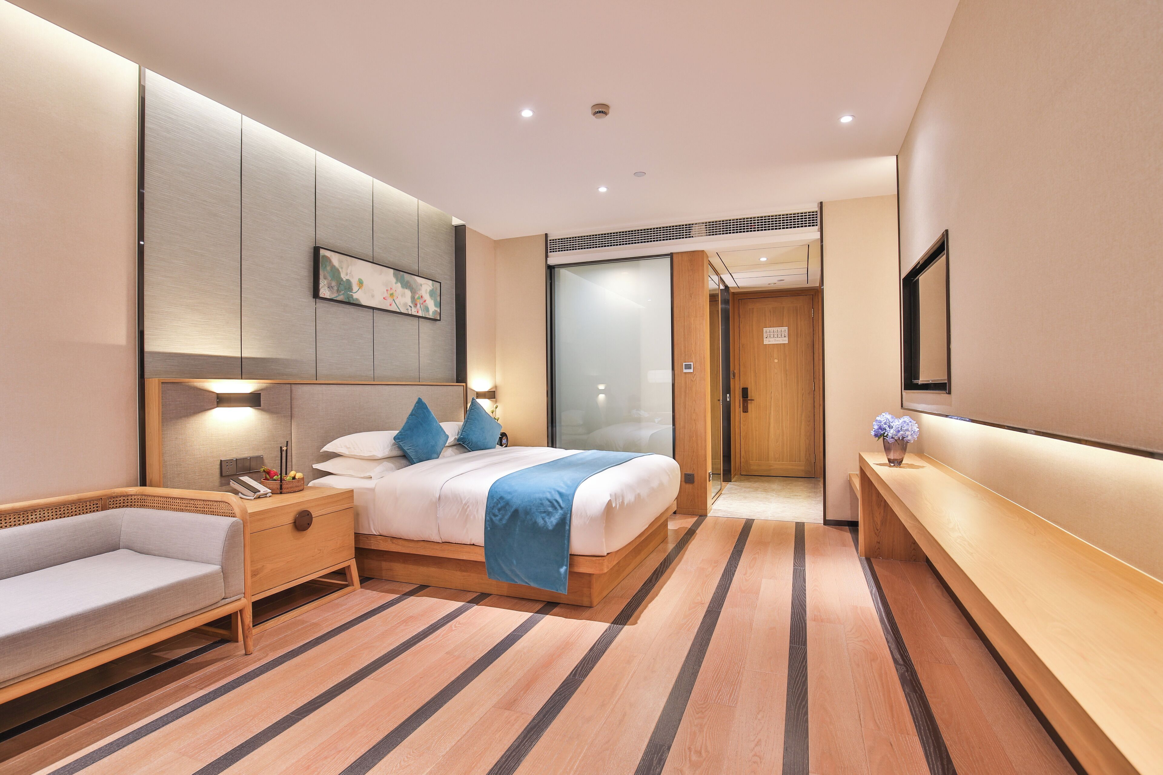 A modern hotel room with a large bed, bedside tables, a sofa, a TV, and a wooden bench.