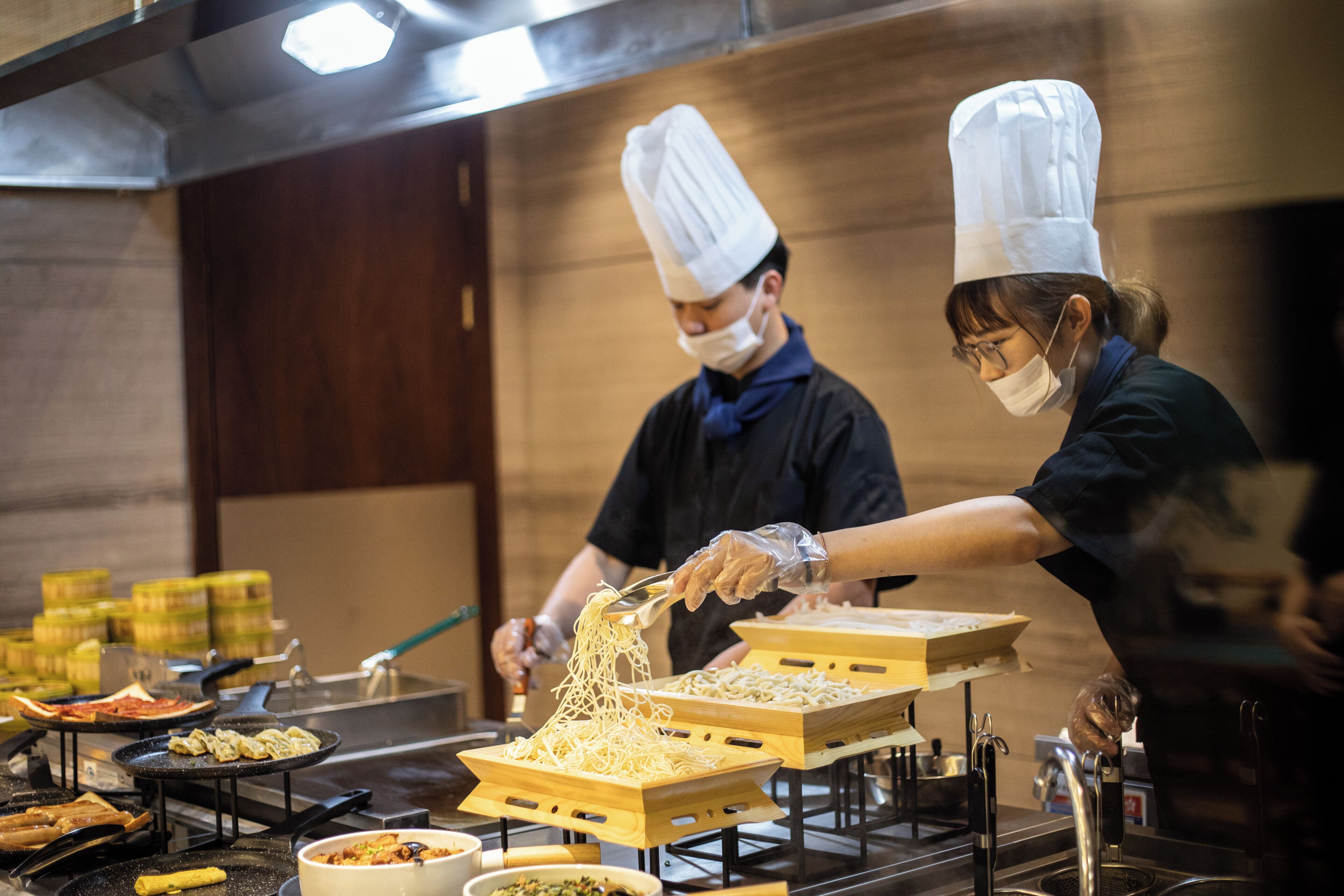 A kitchen with chefs preparing food, including noodles and various dishes on trays.