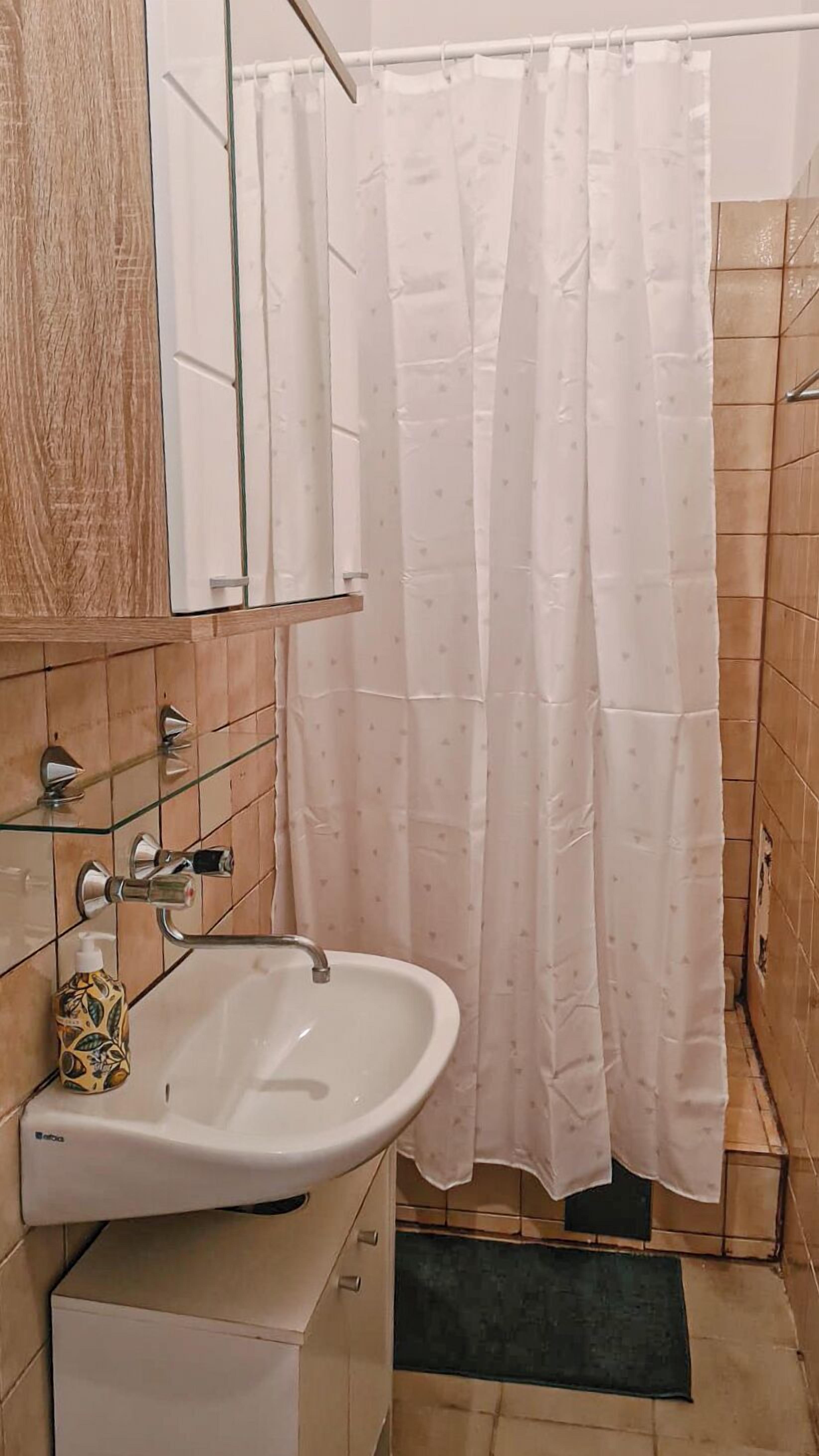 Een badkamer met een wit wastafelmeubel, een glazen plank, een fles handzeep en een douchegordijn.
