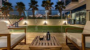Een terras buiten met houten meubels, een kleine tafel en uitzicht op een goed verlicht sportgebied met fitnessapparatuur.