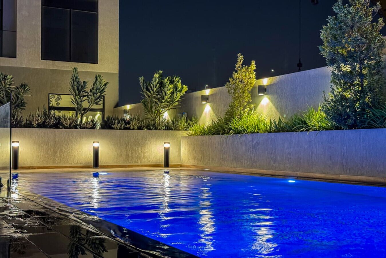 Una piscina nocturna con plantas iluminadas y un edificio moderno al fondo.