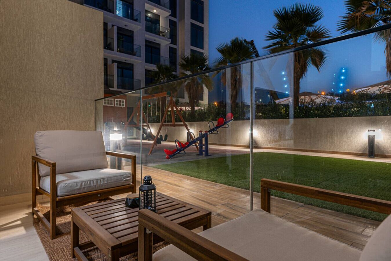 An outdoor balcony with wooden furniture, a swing set, and a view of a cityscape at dusk.