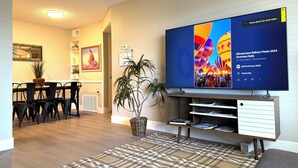 Uma sala de estar moderna com televisão de tela plana exibindo uma festa de balões, uma área de jantar com mesa e cadeiras, e uma planta em vaso.