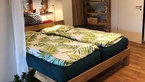 Quarto com cama, mesa de cabeceira, luminária, janela com persianas, prateleira com objetos decorativos e porta.