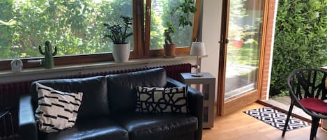 Una sala de estar moderna con un sofá de cuero negro, dos plantas en macetas, una lámpara y vistas al verdor del exterior.