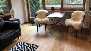 Uma sala de estar moderna com piso de madeira, um sofá de couro preto, uma mesa de jantar com duas cadeiras e janelas amplas com vista para áreas verdes e uma cerca de madeira.