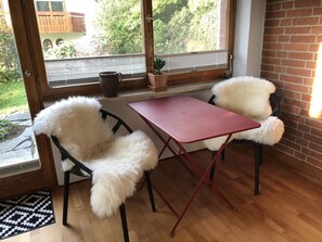 Una habitación con piso de madera, una mesa roja, dos sillas blancas y mullidas, una ventana con persianas y vista a la vegetación exterior.