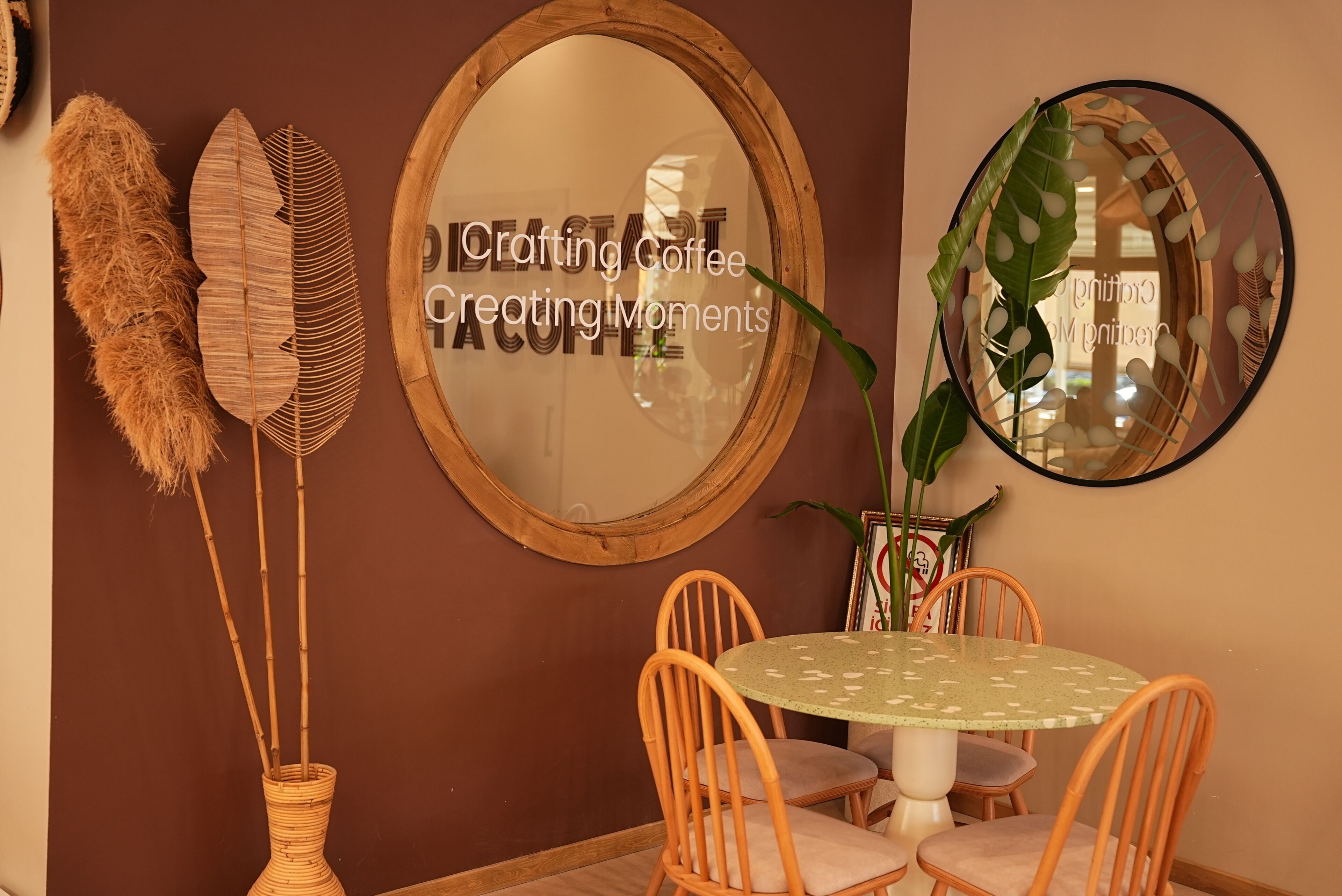 Un comedor con una mesa redonda, sillas de mimbre, un espejo y plantas decorativas.
