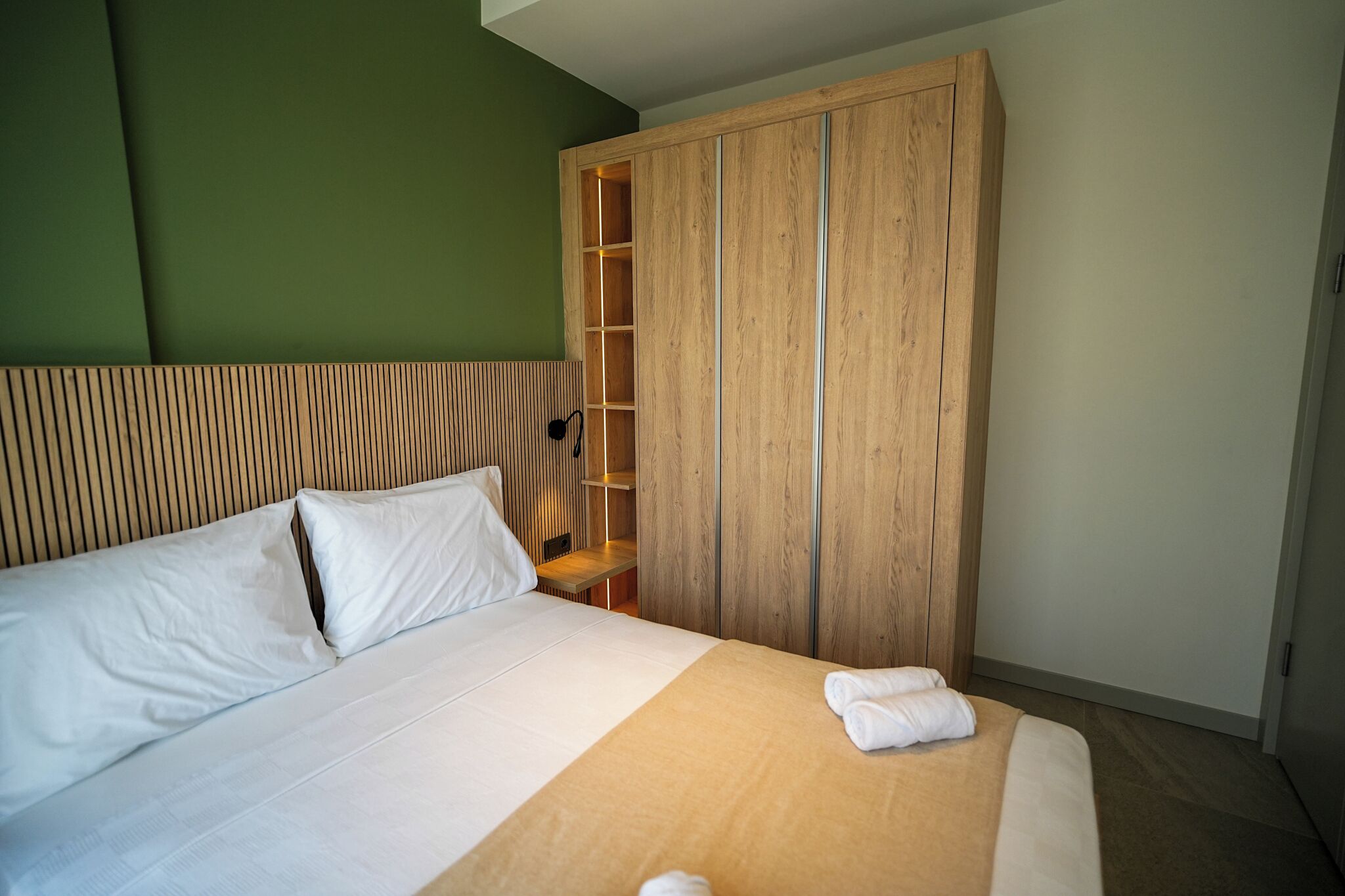 Uma cama bem arrumada com roupa de cama branca, um guarda-roupa de madeira com prateleira embutida e uma parede verde como destaque.