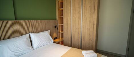 Uma cama bem arrumada com roupa de cama branca, um guarda-roupa de madeira com prateleira embutida e uma parede verde como destaque.
