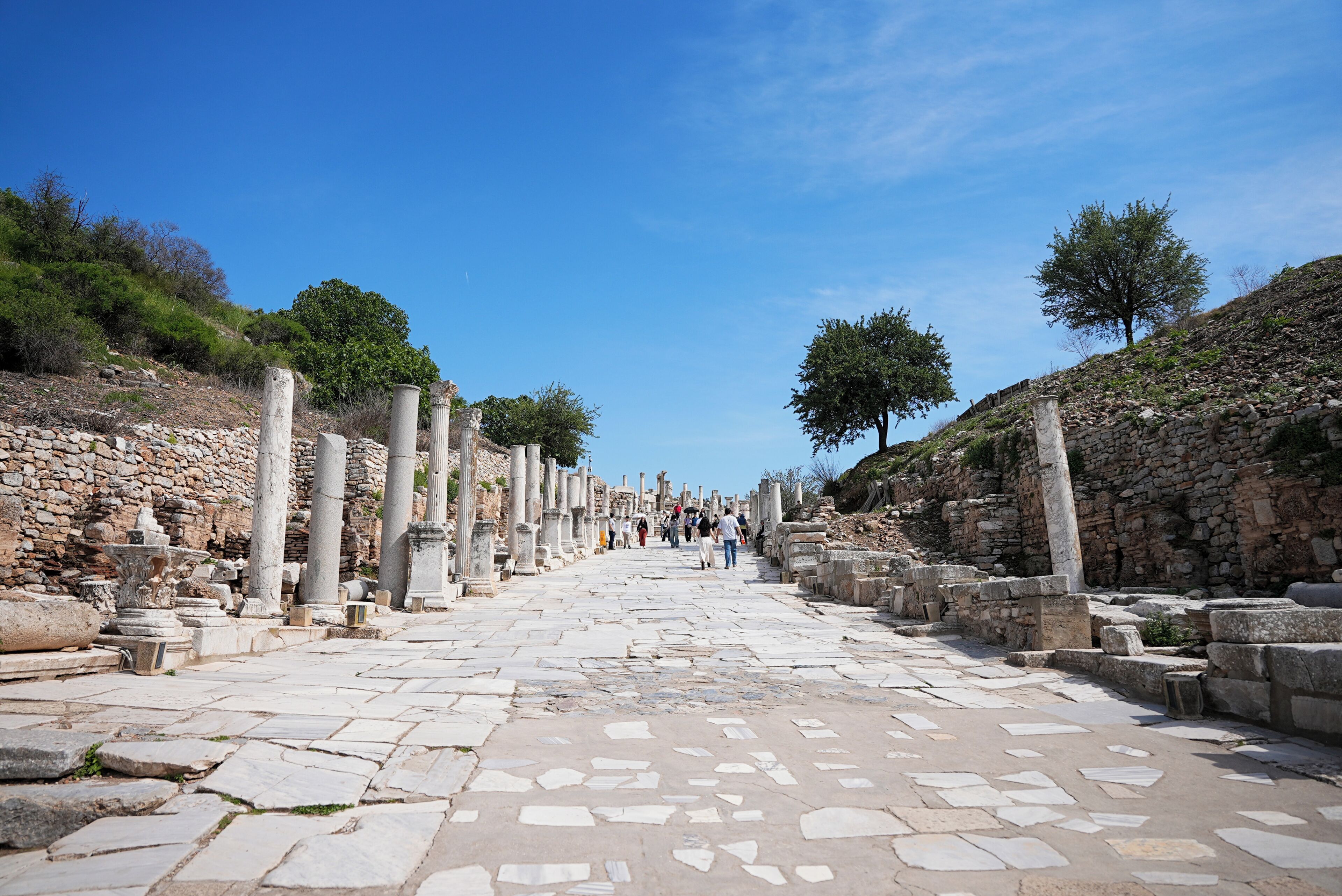 Un ancien sentier pavé de pierres, bordé de colonnes et de ruines, qui mène vers un groupe de personnes se trouvant au loin.