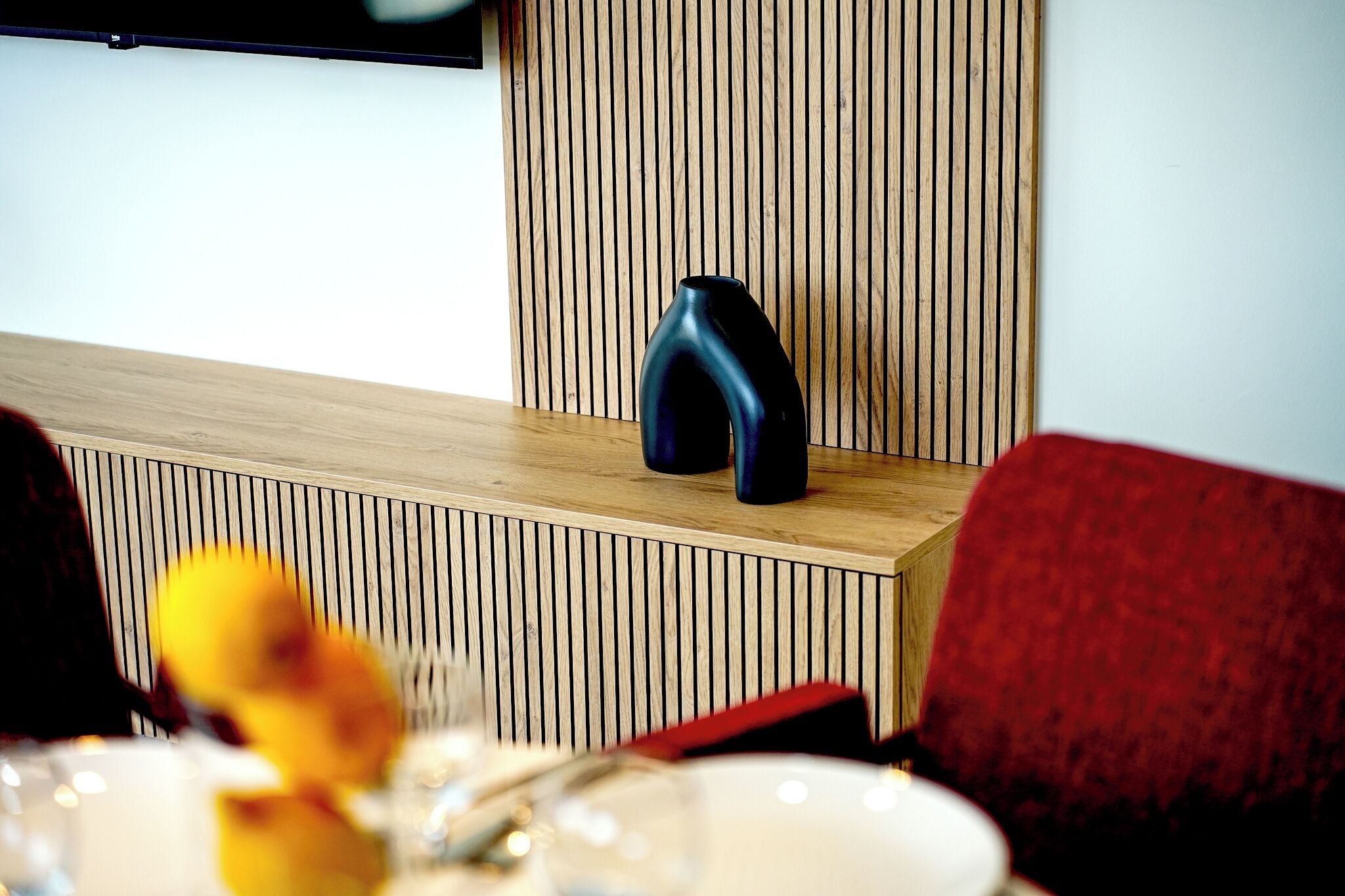 A modern interior with a wooden slatted wall, a black vase on a wooden shelf, and a red upholstered chair.