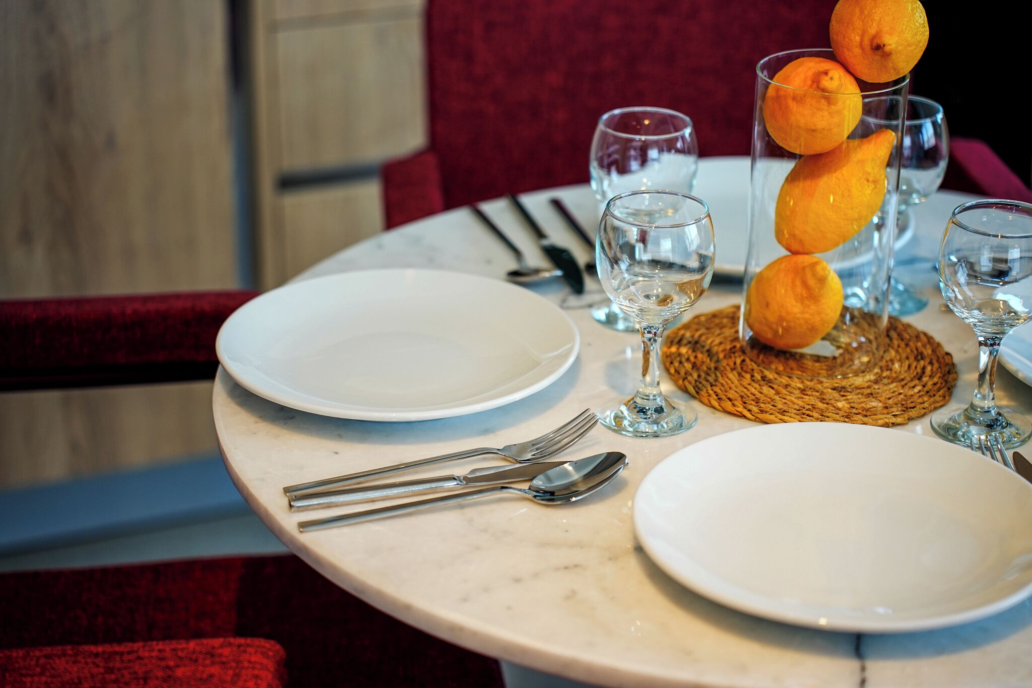 A dining table set with white plates, cutlery, and glasses, with a woven placemat and a stack of oranges.