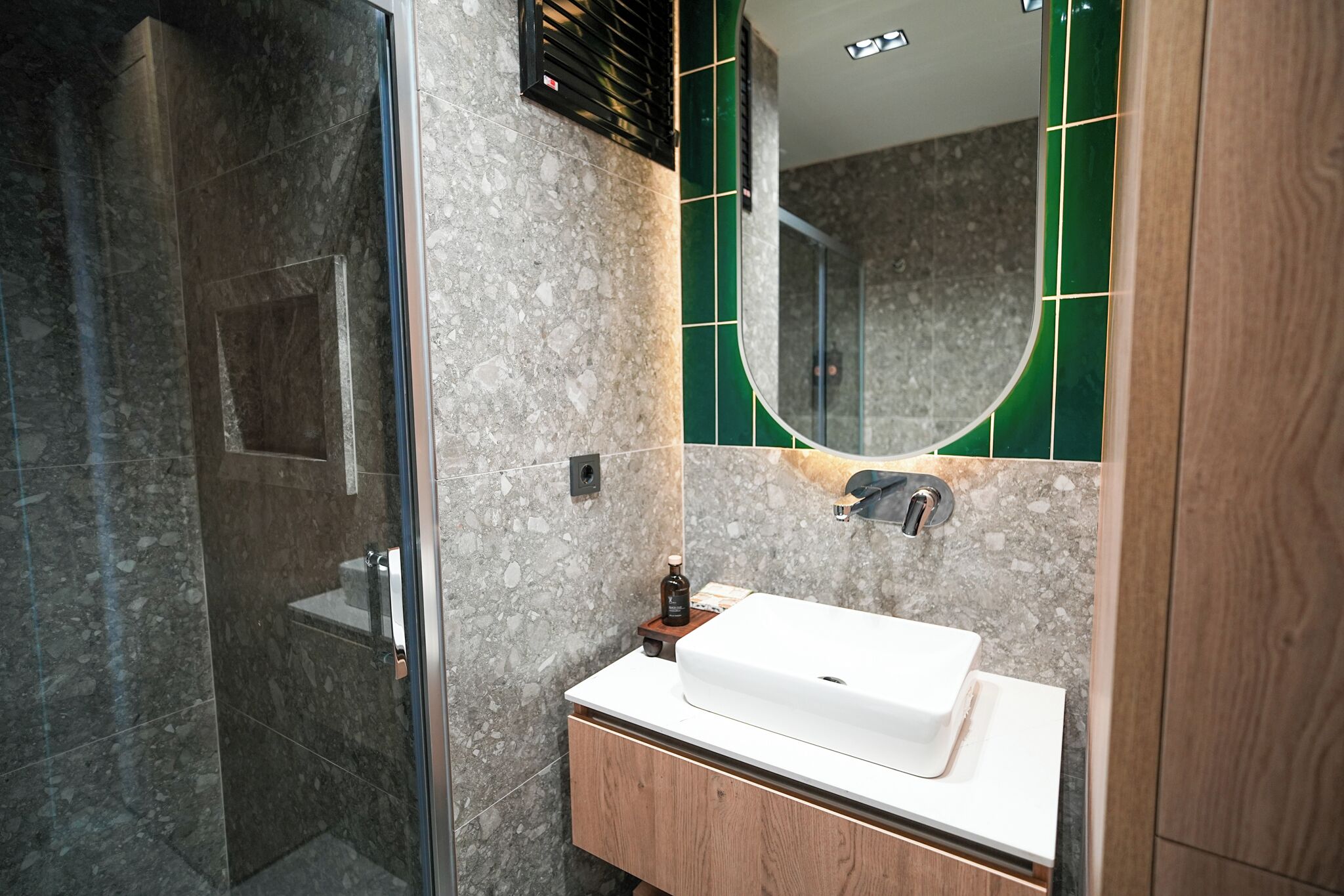 Ein modernes Badezimmer mit Duschkabine aus Glas, einem weißen Waschbecken und einem Spiegel mit grünen Fliesen.