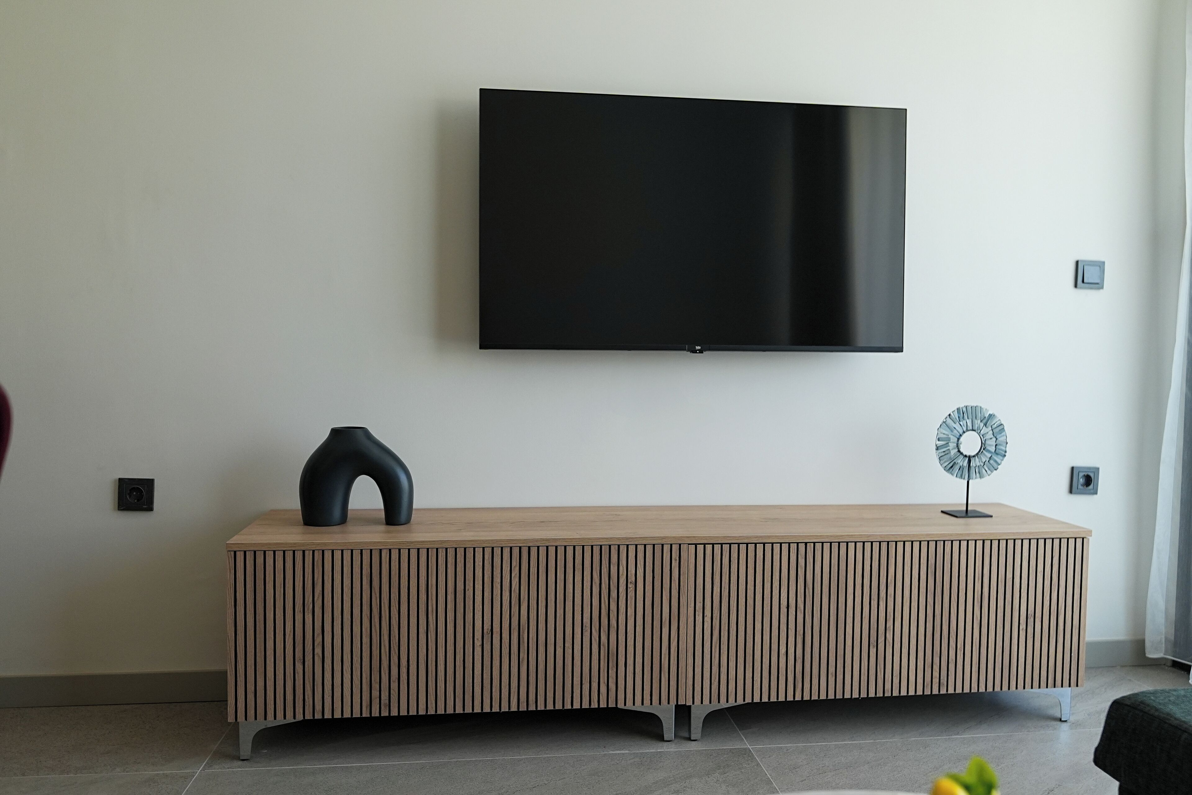 Een moderne woonkamer met een flatscreen televisie aan de muur, een houten console tafel met verticale lamellen, en een decoratieve ventilator.