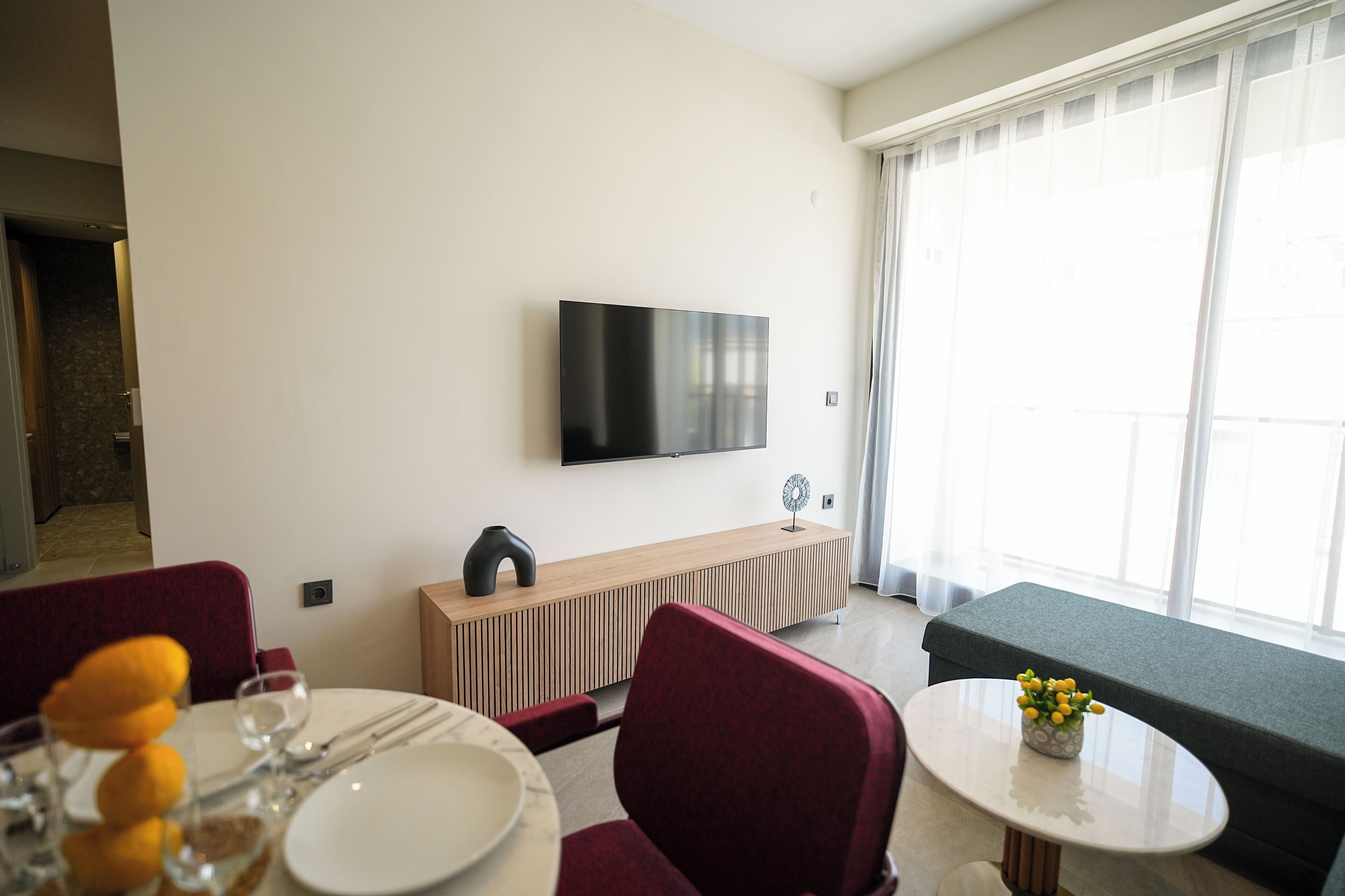 A modern living room with a flat-screen TV mounted on the wall, a wooden console table, a round dining table with a vase of flowers, red upholstered chairs, and a grey sofa.