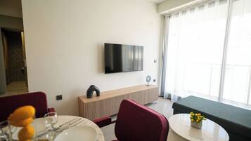 A modern living room with a flat-screen TV mounted on the wall, a wooden console table, a round dining table with a vase of flowers, red upholstered chairs, and a grey sofa.